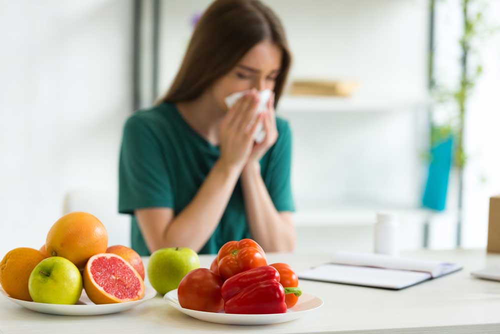Sick woman blowing nose behind plates of fruits and vegetables